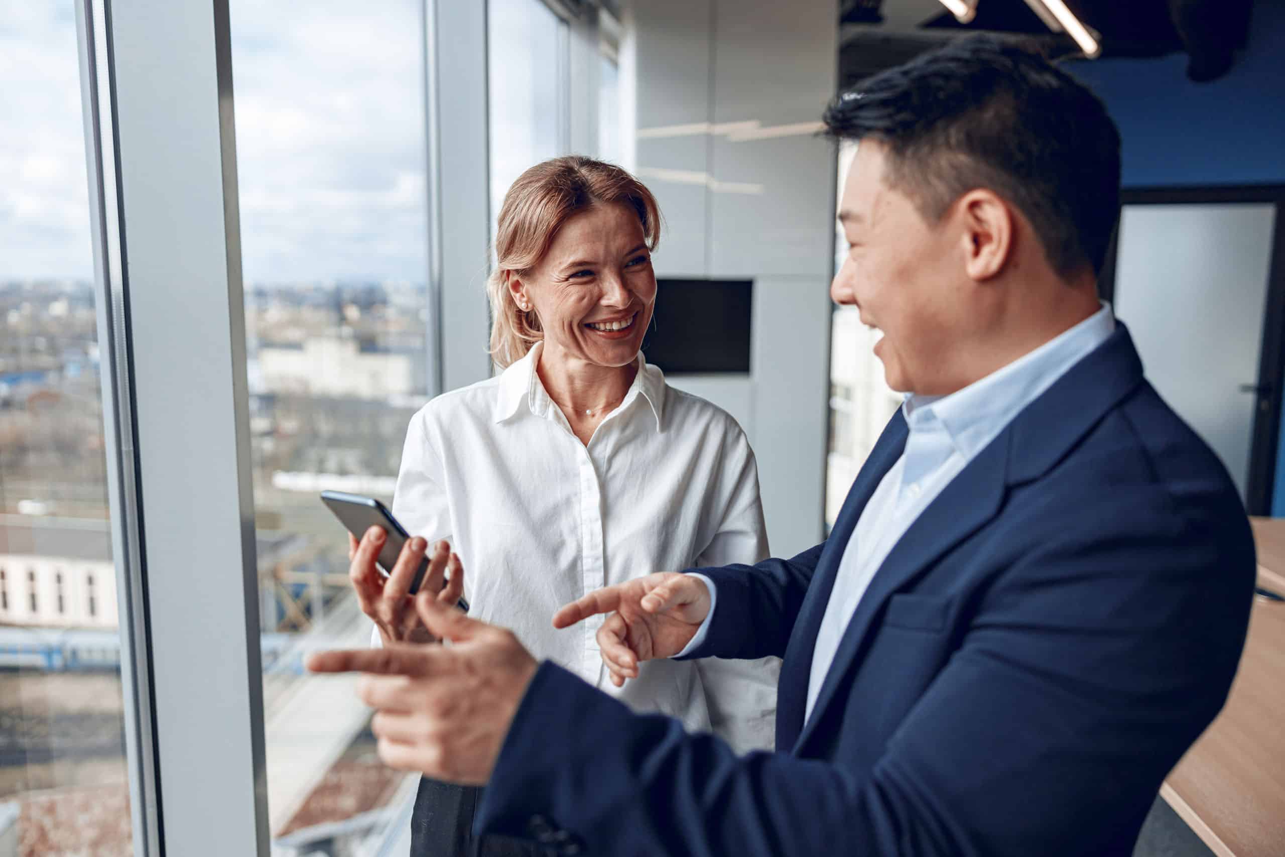 Asiatischer Geschäftsmann unterhält sich selbstbewusst mit einer Geschäftspartnerin vor einem großen Panoramafenster