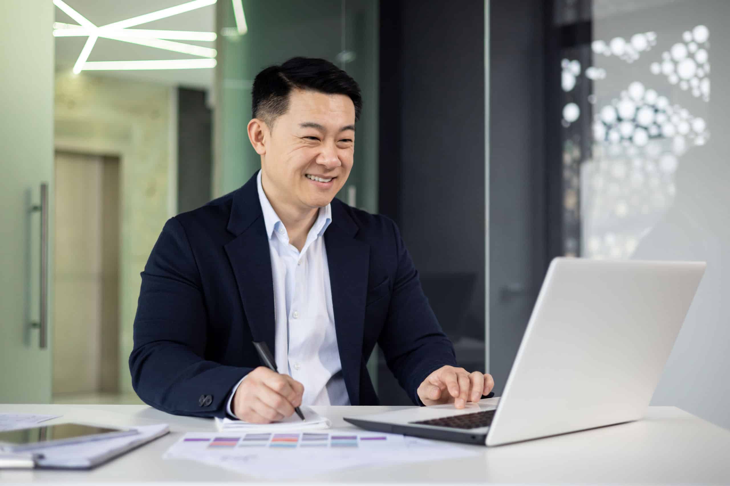 Fröhlicher asiatischer Geschäftsmann im Anzug, der in einem modernen Büro an einem Laptop arbeitet und Notizen macht, dabei Selbstbewusstsein und Produktivität zeigt