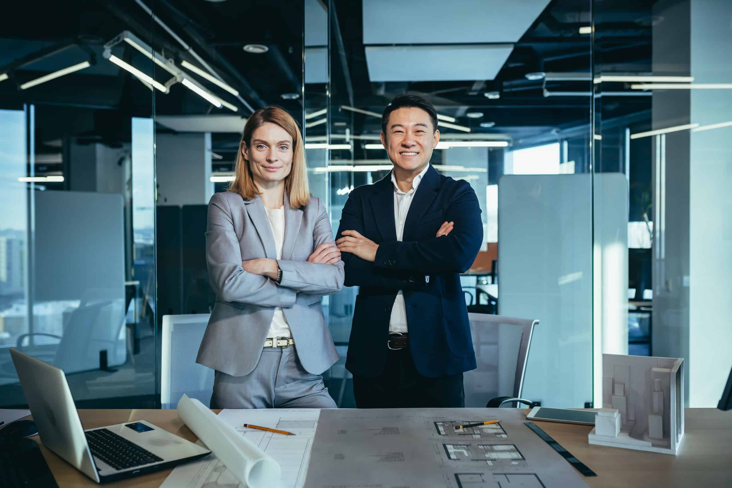 Zwei erfolgreiche Geschäftspartner – ein asiatischer Mann und eine europäische Frau – stehen mit verschränkten Armen in einem modernen Büro