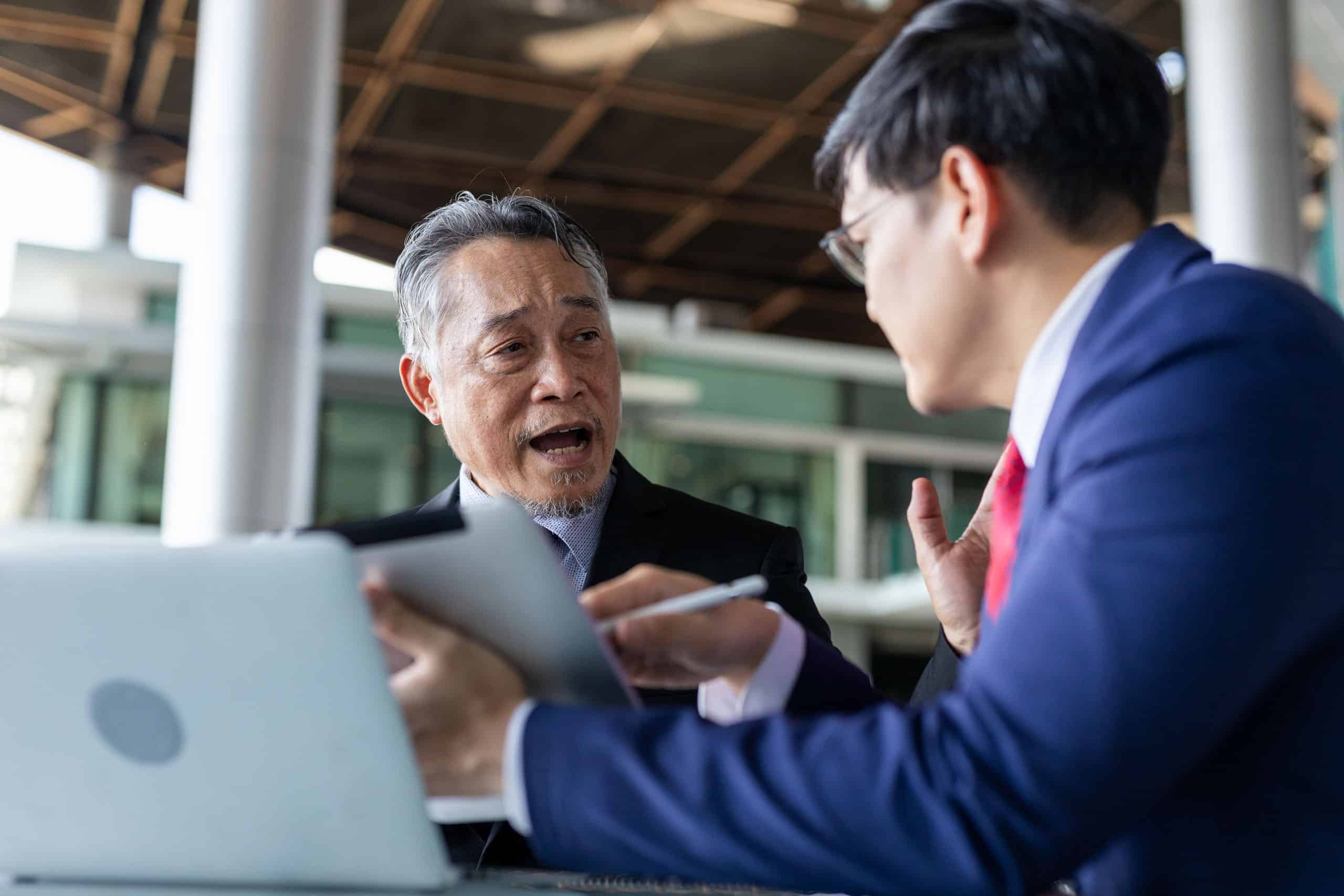 Älterer und jüngerer Geschäftsmann besprechen eine Strategie mithilfe von einem Tablet und einem Laptop