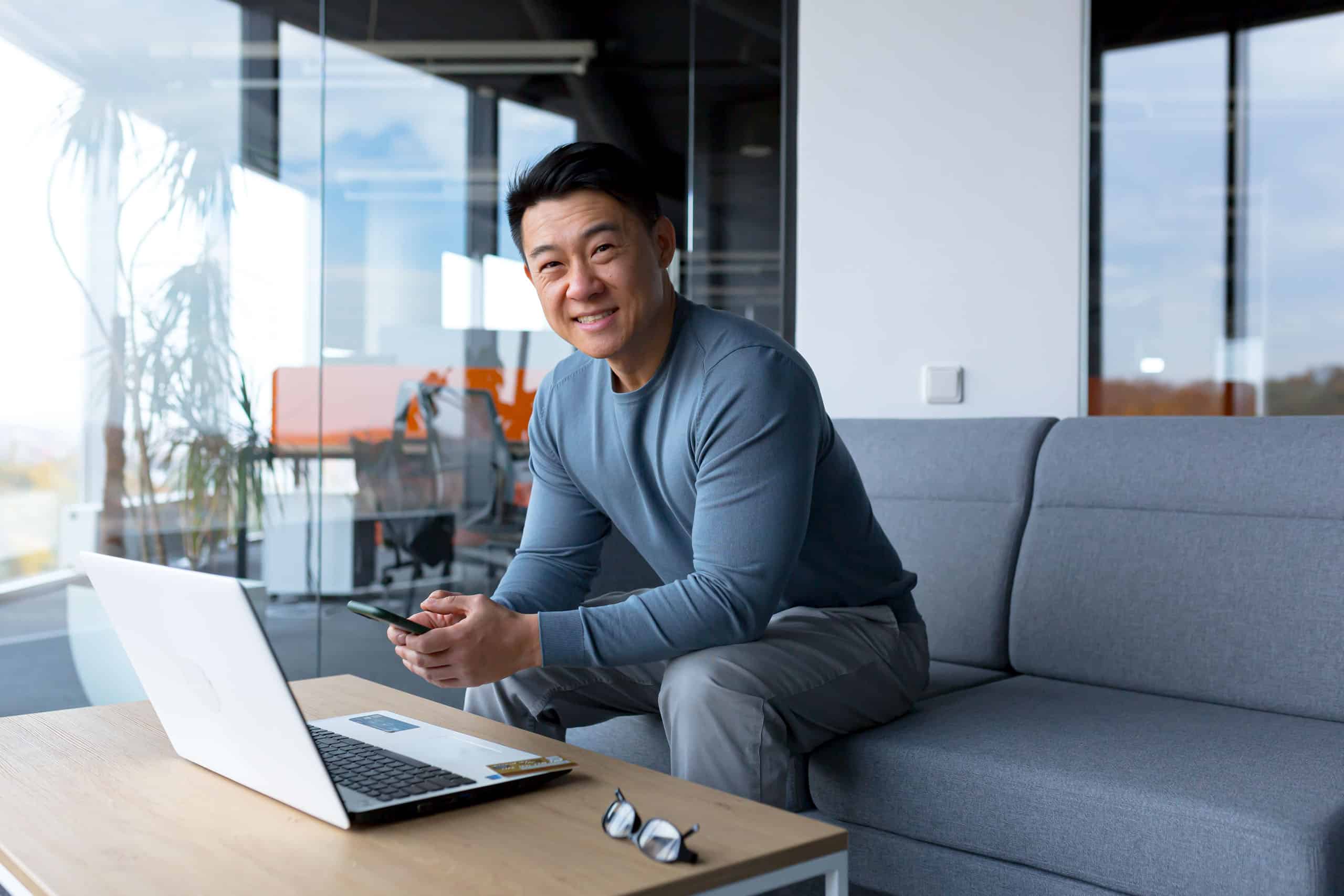 Glücklicher und erfolgreicher asiatischer Geschäftsmann arbeitet mit Laptop auf einem Sofa, schaut lächelnd in die Kamera und hält ein Telefon in der Hand